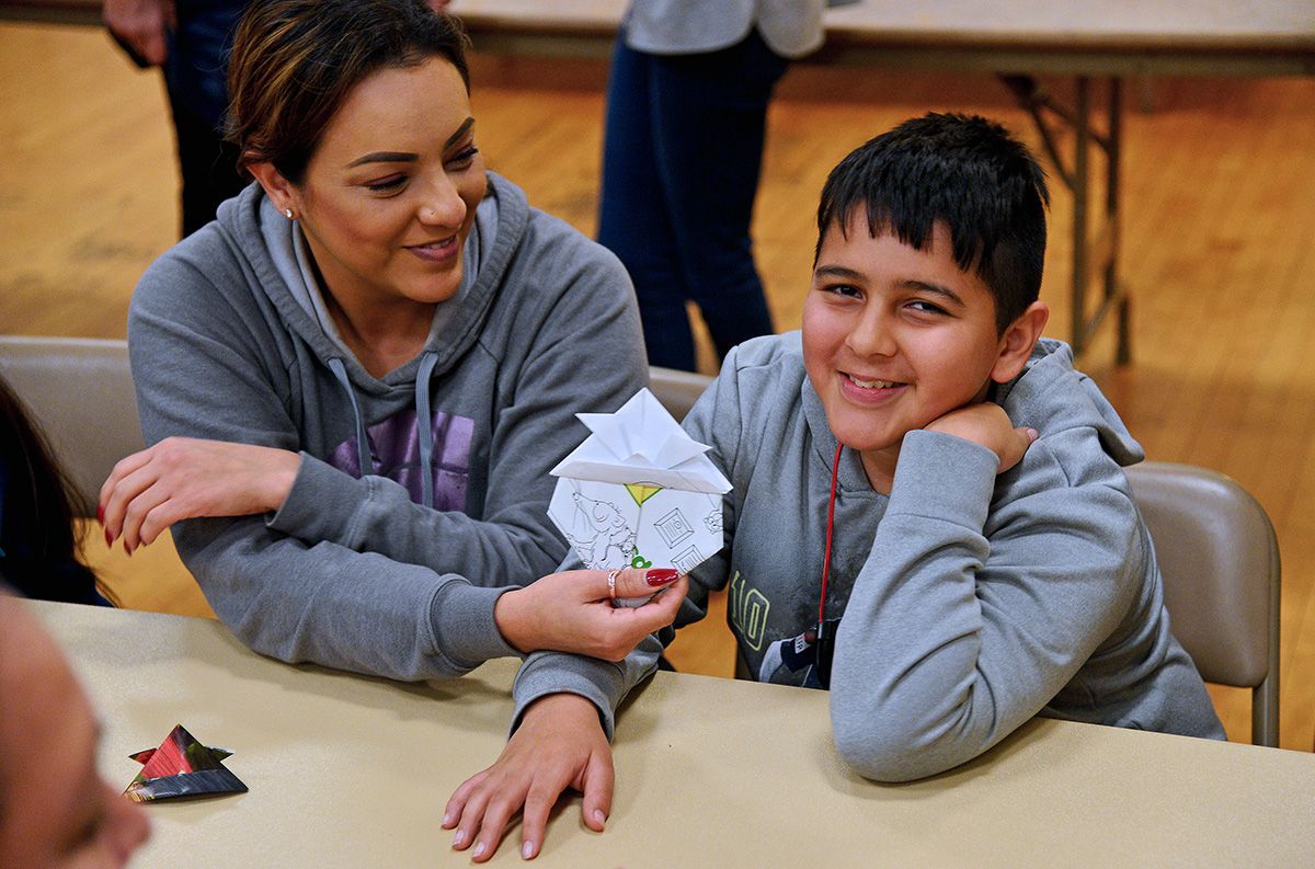 a smiling adult woman holding a geometric paper sculpture made by the smiling boy sitting next to her